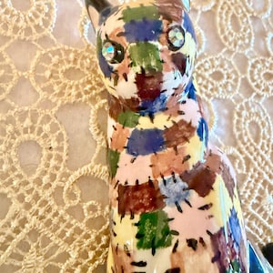May include: A ceramic cat figurine with a patchwork design in various colors, including blue, green, brown, and pink. The cat has large, sparkling eyes and black ears. The figurine is set against a cream-colored, lace-like background.