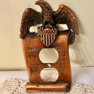 Puede incluir: Cubierta decorativa de enchufe de color cobre con un águila y un pergamino. El pergamino tiene texto y dos aberturas para enchufes. El diseño añade un toque patriótico.