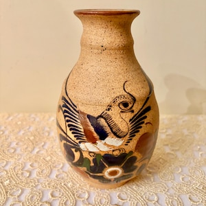 May include: A tan ceramic vase with a bird design. The vase has a speckled texture and a flared rim. The bird is depicted with blue, white, and brown wings, and is surrounded by floral motifs.