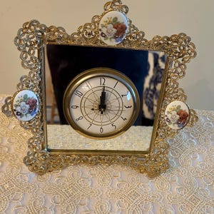 May include: A vintage gold-toned clock with a square mirror face. The clock face is white with black numerals and hands. The frame is ornate with floral accents and decorative ceramic pieces. The clock is set on a lace tablecloth.