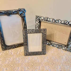 May include: Three silver-colored picture frames of varying sizes. The largest frame has rose and leaf detailing. The medium frame has a decorative scroll design. The smallest frame has a square shape with a detailed border.