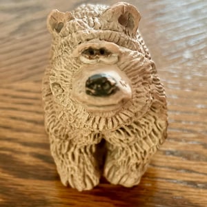 May include: A small, light brown bear figurine with a textured, carved appearance. The bear has a prominent snout, a dark black nose, and detailed fur markings across its body. It is depicted in a seated position, facing forward with a neutral expression.