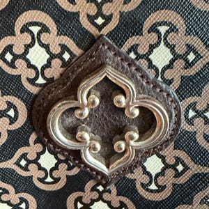 May include: Close-up of a handbag with a repeating pattern in black, beige, and brown. The bag features a dark brown leather accent with a silver-colored decorative emblem in the center.