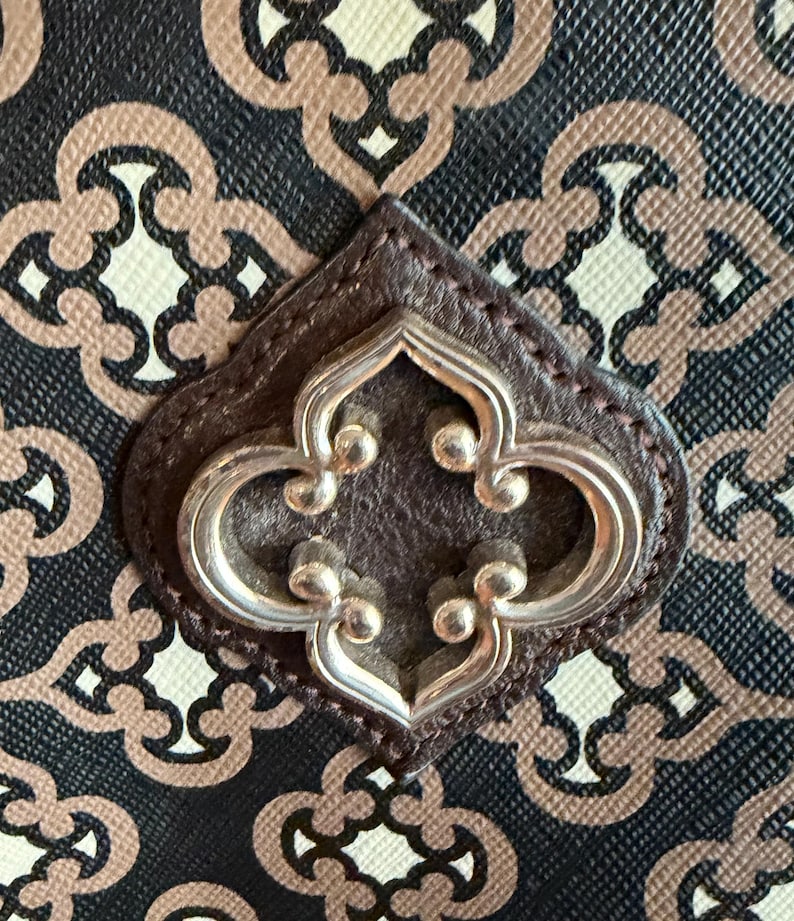May include: Close-up of a handbag with a repeating pattern in black, beige, and brown. The bag features a dark brown leather accent with a silver-colored decorative emblem in the center.