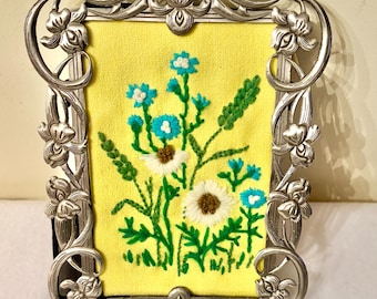 Può includere: Una cornice rettangolare in metallo color argento con dettagli floreali in rilievo, contenente un tessuto giallo ricamato con un bouquet di margherite bianche, fiordalisi blu brillante e steli di grano verdi.