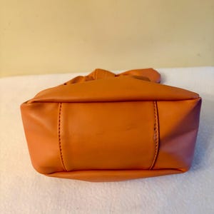 K&ouml;nnte beinhalten: Eine orangefarbene Handtasche mit geraffter Oberseite und rechteckiger Basis. Die Tasche besteht aus einem glatten Material und weist sichtbare N&auml;hte entlang der N&auml;hte auf. Die Farbe der Tasche ist ein leuchtendes, warmes Orange.