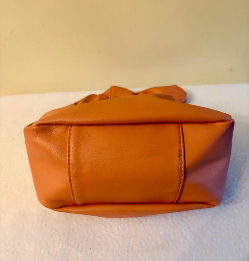 K&ouml;nnte beinhalten: Eine orangefarbene Handtasche mit geraffter Oberseite und rechteckiger Basis. Die Tasche besteht aus einem glatten Material und weist sichtbare N&auml;hte entlang der N&auml;hte auf. Die Farbe der Tasche ist ein leuchtendes, warmes Orange.