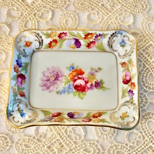 Könnte beinhalten: Rechteckige Porzellanablage mit floralem Design. Die Ablage hat eine weiße Basis mit einem farbenfrohen Blumenarrangement in der Mitte und an den Rändern. Goldener Rand.