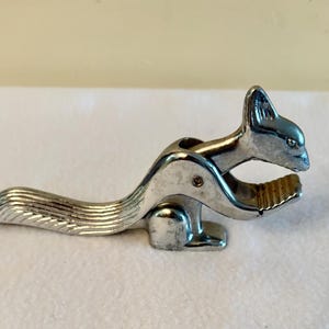 May include: A silver-colored metal nutcracker shaped like a squirrel. The tool features a long, textured tail that serves as a handle, a hinged body, and a serrated jaw area designed for cracking nuts. The metallic surface has a polished finish.