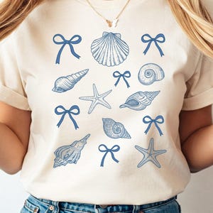 May include: A white t-shirt with a blue seashell and starfish design. The design includes blue bows and seashells in various shapes.