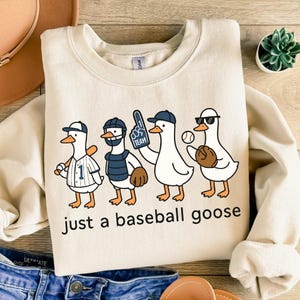May include: Cream-colored sweatshirt featuring a cartoon illustration of four geese dressed as baseball players. The geese are wearing baseball caps, mitts, and uniforms. The text "just a baseball goose" is printed below the illustration.