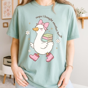 Op de afbeelding: Lichtblauw T-shirt met een cartoon gans met een roze strik, laarzen en boeken. De tekst "oops i bought another book" staat boven de gans. Witte bloemen en gouden sterren versieren het shirt.