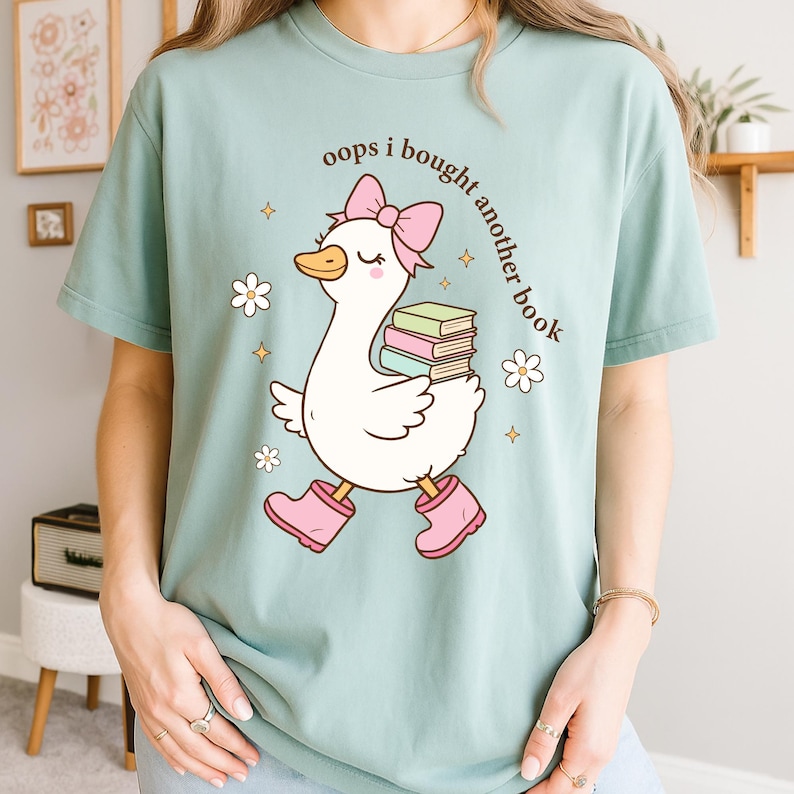 Op de afbeelding: Lichtblauw T-shirt met een cartoon gans met een roze strik, laarzen en boeken. De tekst "oops i bought another book" staat boven de gans. Witte bloemen en gouden sterren versieren het shirt.