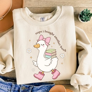 Op de afbeelding: Cr&egrave;mekleurige sweatshirt met een grillige afbeelding van een witte gans met een roze strik, laarzen en boeken. De tekst "oops i bought another book" staat boven de gans gedrukt. Het ontwerp bevat bloemen en sterren.