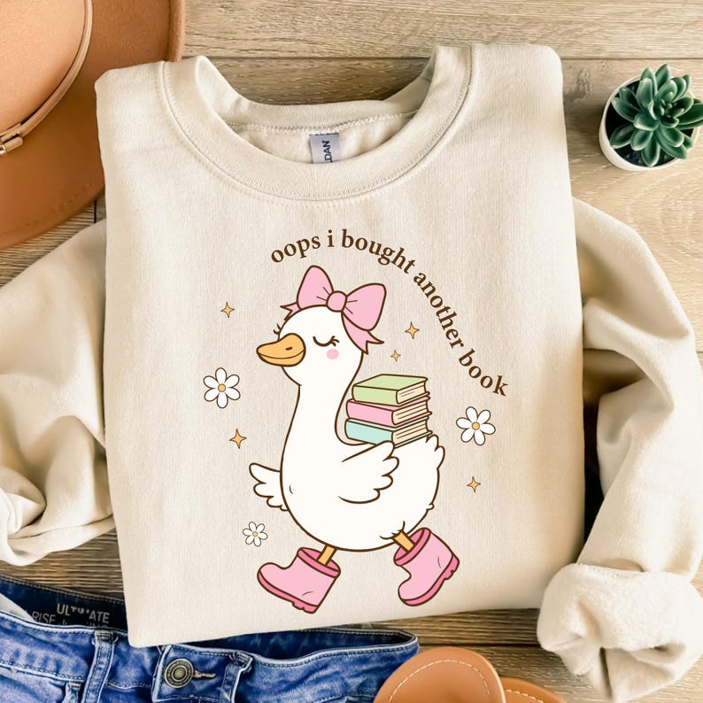 Op de afbeelding: Cr&egrave;mekleurige sweatshirt met een grillige afbeelding van een witte gans met een roze strik, laarzen en boeken. De tekst "oops i bought another book" staat boven de gans gedrukt. Het ontwerp bevat bloemen en sterren.