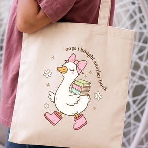 Op de afbeelding: Cr&egrave;mekleurige draagtas met een cartoon gans met een roze strik, laarzen en boeken. De tekst "oops i bought another book" staat op de tas. Het ontwerp bevat bloemen en sterren.