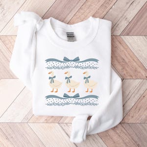May include: White sweatshirt featuring a blue and white design of three geese with blue bows. The geese are arranged in a row with a blue ribbon and polka dots above them.
