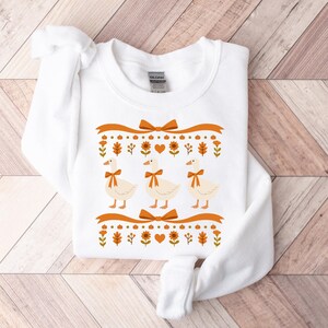 May include: White sweatshirt featuring three geese with orange bow ties, set against a decorative orange ribbon and fall-themed border. The border includes pumpkins, leaves, and flowers. The design is centered on the front of the sweatshirt.