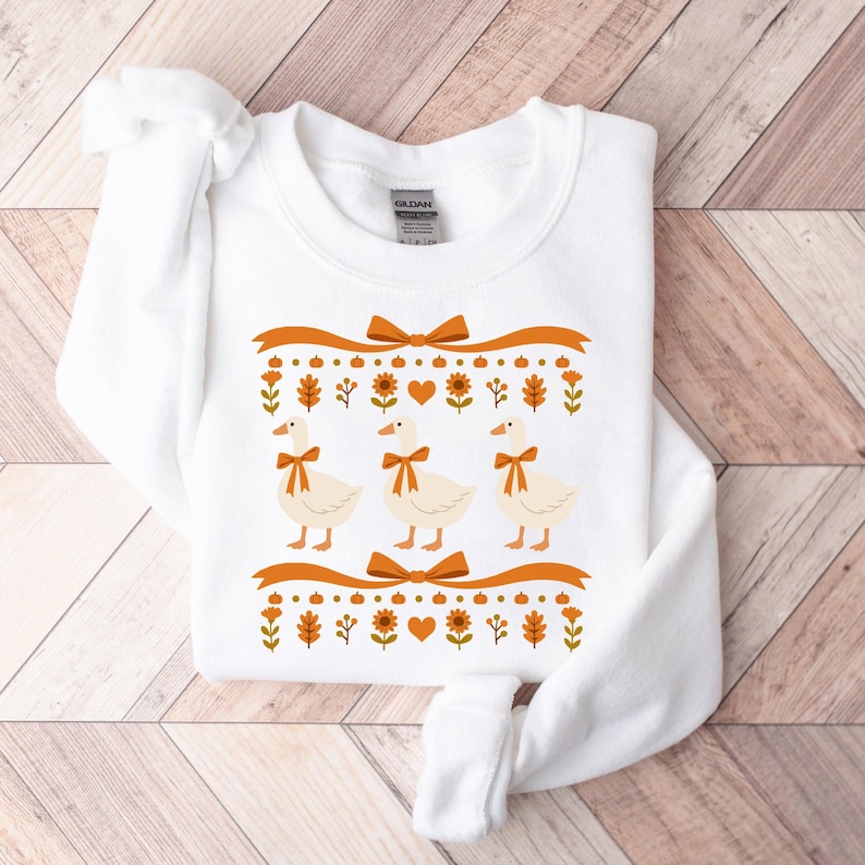 May include: White sweatshirt featuring three geese with orange bow ties, set against a decorative orange ribbon and fall-themed border. The border includes pumpkins, leaves, and flowers. The design is centered on the front of the sweatshirt.