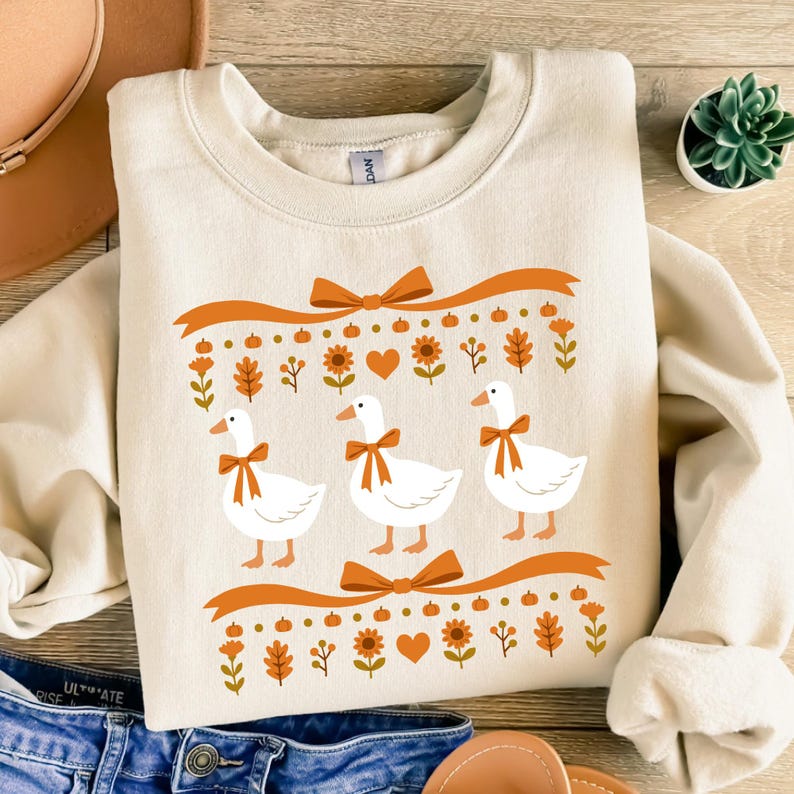 May include: Cream-colored sweatshirt featuring a whimsical design of three white geese with orange bow ties. The design includes orange ribbons, pumpkins, flowers, and hearts, creating a fall-themed aesthetic.