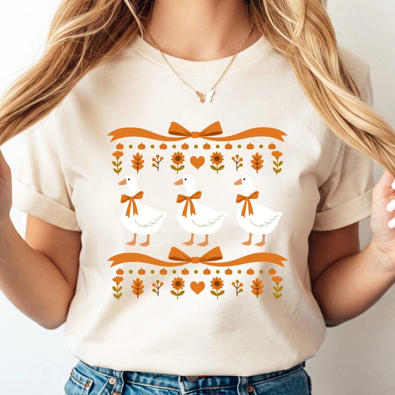 May include: Cream-colored t-shirt featuring three white geese with orange bow ties. The design includes orange ribbons, bows, and fall-themed floral and leaf elements. A gold necklace with a butterfly pendant is visible.