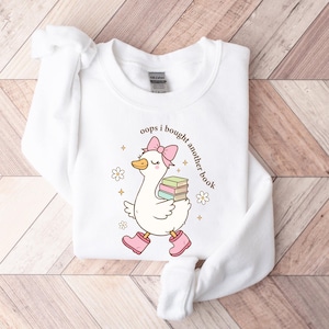 Op de afbeelding: Witte sweatshirt met een cartoon eend met roze laarzen en een strik, met boeken. De tekst "oops i bought another book" staat op de sweatshirt. Het ontwerp bevat bloemen en sterren.