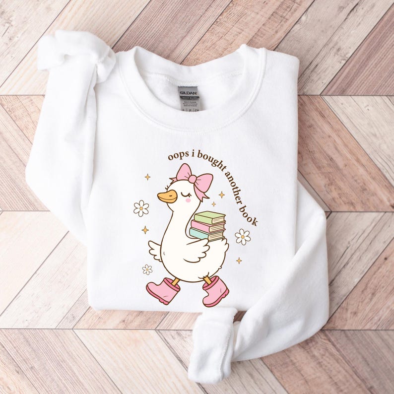 Op de afbeelding: Witte sweatshirt met een cartoon eend met roze laarzen en een strik, met boeken. De tekst "oops i bought another book" staat op de sweatshirt. Het ontwerp bevat bloemen en sterren.