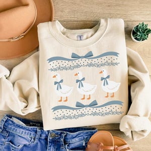 May include: A cream-coloured sweatshirt with a blue and white pattern of geese and bows. The geese are white with blue bows around their necks. The pattern is repeated twice on the sweatshirt.