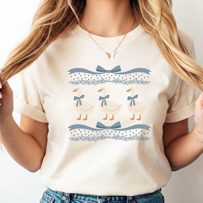 May include: A white t-shirt featuring a blue and white pattern of geese, bows, and leaves. The geese are wearing blue bows.