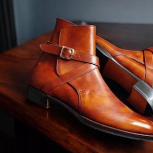 May include: A pair of brown leather ankle boots with a pointed toe and a strap with a gold buckle. The boots are on a wooden surface. The boots have a classic design.