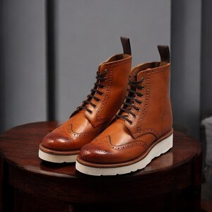 Pu&ograve; includere: Un paio di stivali in pelle marrone con lacci neri e suole bianche. Gli stivali hanno un classico design a punta alata con perforazioni decorative. Gli stivali sono appoggiati su una superficie di legno scuro.