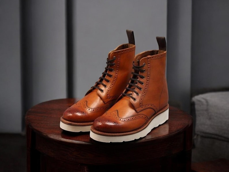 Pu&ograve; includere: Un paio di stivali in pelle marrone con lacci neri e suole bianche. Gli stivali hanno un classico design a punta alata con perforazioni decorative. Gli stivali sono appoggiati su una superficie di legno scuro.