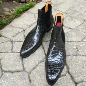 May include: A pair of dark green Chelsea boots with a glossy, crocodile-skin texture. The boots have a pointed toe and elastic side panels. The interior lining is visible, with a red accent. The boots are displayed on a stone surface.