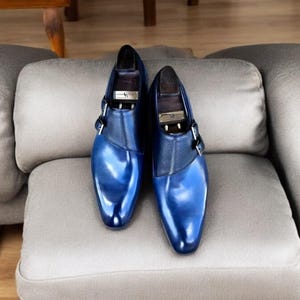 以下が含まれることがあります： バックル留めの青い革製のドレスシューズのペア。シューズは薄い灰色のソファの上に置かれています。
