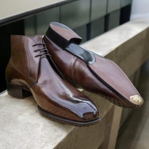 May include: A pair of brown leather dress shoes. One is a lace-up ankle boot, the other a slip-on with a black accent and a gold-colored toe cap. The shoes have a high-gloss finish and are displayed on a stone surface.