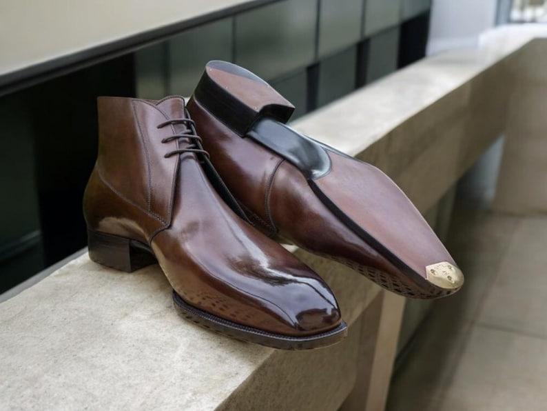 May include: A pair of brown leather dress shoes. One is a lace-up ankle boot, the other a slip-on with a black accent and a gold-colored toe cap. The shoes have a high-gloss finish and are displayed on a stone surface.