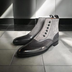 Puede incluir: Un par de botines de cuero gris y negro con cierre de botones y detalles brogue. Los botines tienen un aspecto elegante y pulido, con una combinación de texturas lisas y de ante. Los botines se exhiben sobre una superficie embaldosada.
