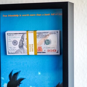 Könnte beinhalten: Gerahmtes Kunstwerk mit einem Stapel von 10.000 US-Dollar-Banknoten, gesichert mit einem gelben Band. Der Hintergrund ist ein blauer Himmel mit Cartoon-Figuren. Der Text oben lautet: „Wahre Freundschaft ist mehr wert als eine Bank voller mo.“