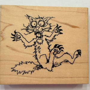 May include: A wooden stamp featuring a cartoon cat with a surprised expression. The cat is drawn in black ink, with wide eyes, an open mouth, and outstretched paws. The stamp is square and made of light-colored wood.