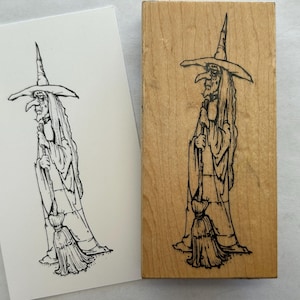 May include: A black and white illustration of a witch with a tall hat and broom. The witch is depicted in a side profile, with long hair and a long nose. The image is on a white card and a wooden stamp.