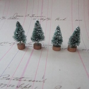Peut inclure: Quatre petits sapins artificiels enneig&eacute;s sur un fond blanc avec des lignes roses et bleues. Les sapins sont verts avec un flocage blanc et sont sur des bases brunes.