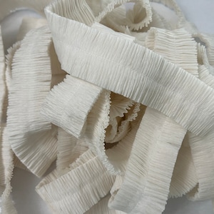 May include: A close-up of a white ruffled trim. The trim is made of a soft, crinkled material and has a delicate, frilly edge.