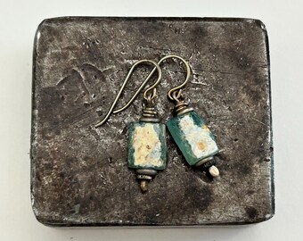 May include: A pair of dangle earrings featuring rectangular, weathered teal-colored glass beads with textured, off-white inclusions. The beads are suspended from dark metal ear wires, secured with coiled wire accents and small metal beads.