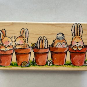 May include: A wooden stamp featuring five cartoon bunnies in terracotta pots. The bunnies have orange and brown fur, and the pots are set on a green grassy base with small flowers and snails. The stamp is a charming illustration.
