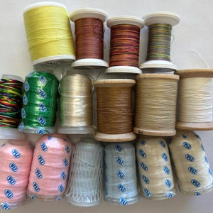 May include: A collection of fifteen spools of thread in various colors including yellow, green, brown, pink, and white. Several spools feature a blue and white YLI logo on the base. The threads are arranged in three rows on a plain surface.