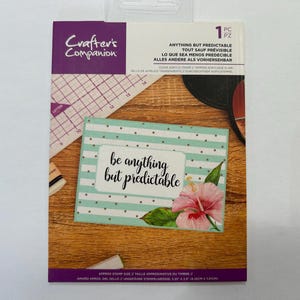 May include: A clear acrylic stamp set by Crafter's Companion. The packaging features the text "Anything But Predictable" in multiple languages. A card with the text "be anything but predictable" and a floral design is also visible.