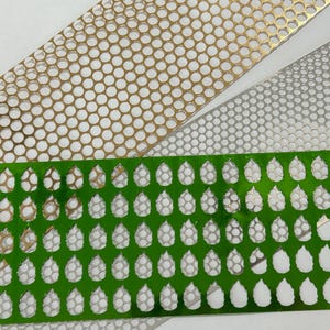 May include: Close-up of three decorative metal strips. One is gold with a circular pattern, another is silver with a similar pattern, and the third is green with a teardrop design. These strips are likely used for crafts or decorative purposes.