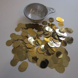 May include: A pile of gold-colored round sequins, some of which are stacked on top of each other, with a silver measuring cup in the background.