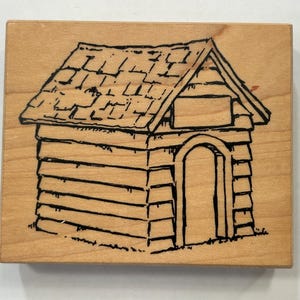 May include: A wooden stamp featuring a detailed black line drawing of a small cabin or house. The cabin has a sloped roof, an arched doorway, and is constructed with horizontal log-like elements. The stamp is on a light-colored wooden block.
