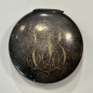 May include: A round, dark, tarnished metal compact case featuring a prominent, intricate gold-toned engraved monogram in the center. The surface shows a mottled, aged patina with a visible hinge at the top edge of the circular container.